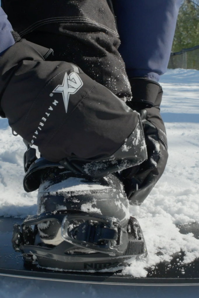 Close-up of a snowboarder strapping in bindings while wearing winter mittens in the snow.